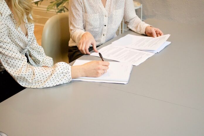 Photo by Gabrielle Henderson woman signing on white printer paper beside woman about to touch the documents