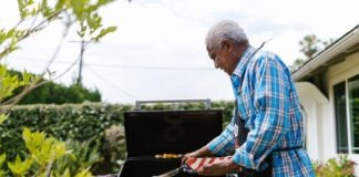 An elderly man grilling sausages on a barbecue in his backyard on a sunny day.
