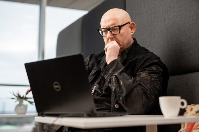 Photo by ODISSEI a bald man sitting in front of a laptop computer
