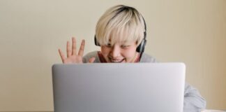 Smiling young Asian female in headphones and casual clothes having conversation via netbook and giving high five while looking at screen and sitting at table with cup of hot drink