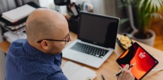 A man in an office setting using a laptop and tablet while taking notes.