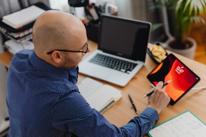 A man in an office setting using a laptop and tablet while taking notes.