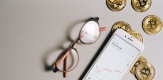 Flat lay of Bitcoin coins, eyeglasses, and smartphone displaying cryptocurrency graph.