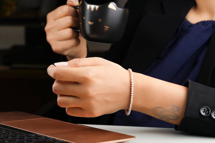 A close-up of a hand wearing a diamond bracelet, holding a cat-themed mug near a laptop.