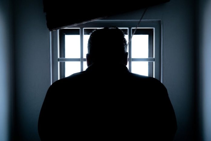 Dark silhouette of a man in a prison cell, backlit by window bars.