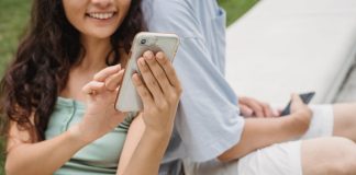 Crop young cheerful female browsing internet on cellphone while resting on bench near unrecognizable partner in town