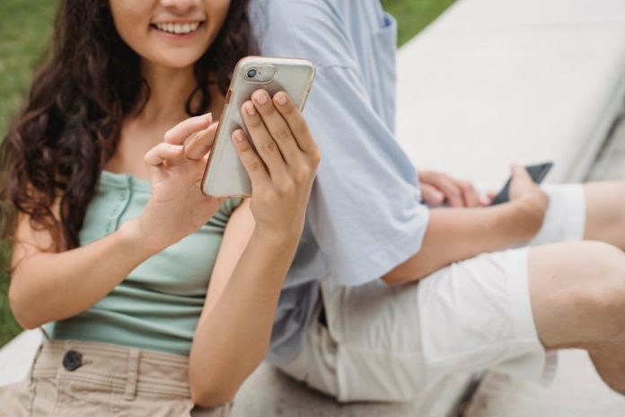 Crop young cheerful female browsing internet on cellphone while resting on bench near unrecognizable partner in town