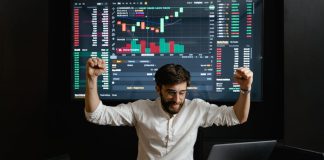 Businessman celebrates stock market success with hands raised in excitement at a trading desk.