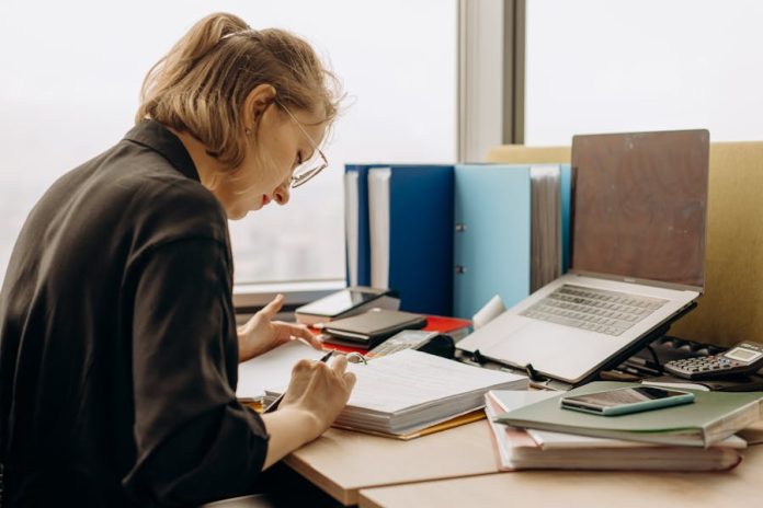 Photo by Mikhail Nilov Woman in glasses intensely working at a desk with documents and a laptop.