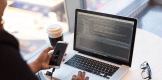 Developer working remotely, coding on a laptop with phone in hand, showcasing modern work culture.