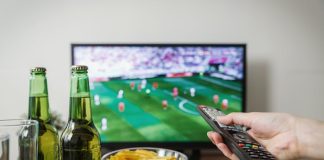 A person holding a remote while watching soccer on TV with beer and snacks on the table.