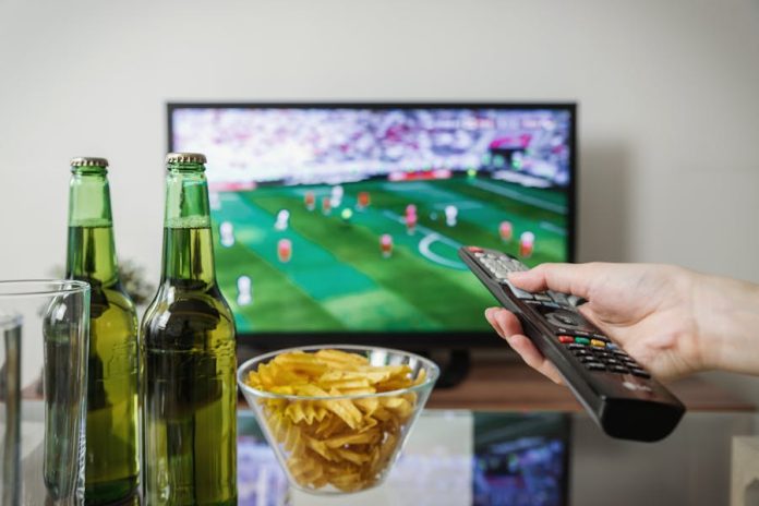 Photo by JESHOOTS.com A person holding a remote while watching soccer on TV with beer and snacks on the table.