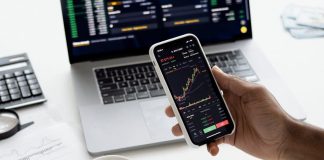 A person trades stocks on a smartphone with a laptop, coffee cup, calculator, and documents in view.