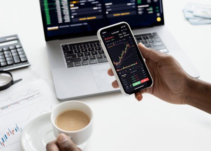 Photo by Anna Nekrashevich A person trades stocks on a smartphone with a laptop, coffee cup, calculator, and documents in view.