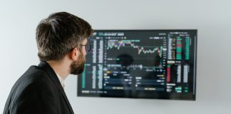 A businessman examines stock market data displayed on a monitor, holding a tablet.