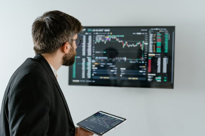Photo by Tima Miroshnichenko A businessman examines stock market data displayed on a monitor, holding a tablet.