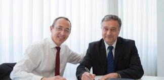 Two businessmen in suits signing a contract at a well-lit office table.