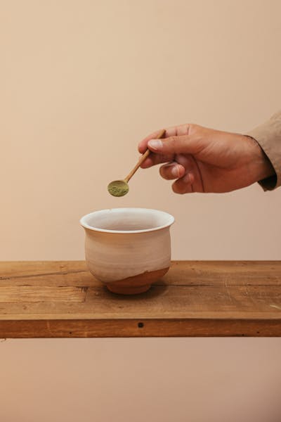 Photo by Cup of Couple A hand carefully adding matcha powder to a ceramic cup positioned on a wooden table.