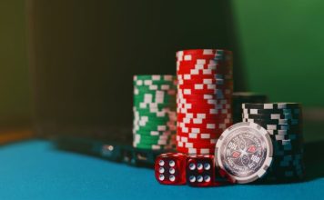 Close-up of casino chips and dice on a felt table, next to a laptop for online gambling.