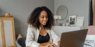 A woman sits at a round table, working on a laptop with a coffee cup nearby in a cozy home office setting.