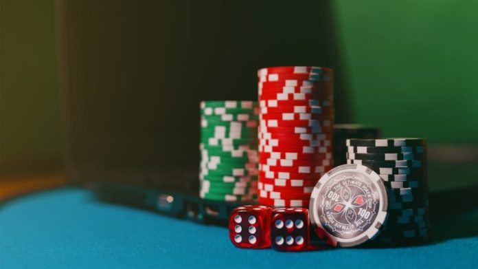 Photo by Aidan Howe Close-up of casino chips and dice on a felt table, next to a laptop for online gambling.