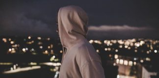 Exclusive Sp5der Hoodie – High Quality Streetwear Fit A person in a hoodie overlooks a blurry cityscape at night, evoking mystery and solitude.