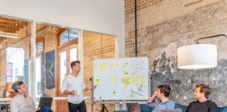 three men sitting while using laptops and watching man beside whiteboard