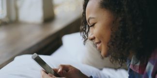 Young woman smiling while using her smartphone in bed.