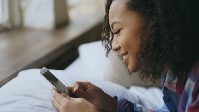 Young woman smiling while using her smartphone in bed.